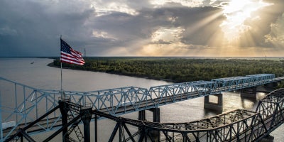 Vicksburg Bridge over the Mississippi River with greenery shown and sun shining in the background as the American flag flies. 