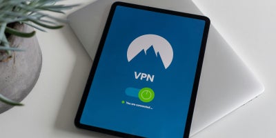 Tablet sitting on top of a laptop computer next to a houseplant displaying “VPN” with a logo of a mountain in the background.