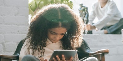 Young girl looking down at a tablet while young boy in the background plays with a toy
