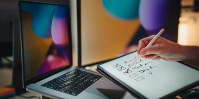 Person sitting in front of a laptop while checking off tasks on a tablet
