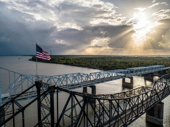 Vicksburg Bridge over the Mississippi River with greenery shown and sun shining in the background as the American flag flies. 
