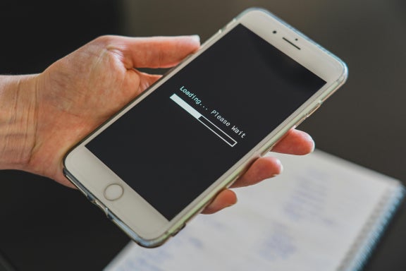 Person holding phone above a notebook displaying “Loading...please wait” as internet is buffering 