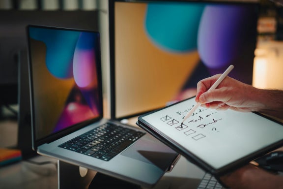 Person sitting in front of a laptop while checking off tasks on a tablet