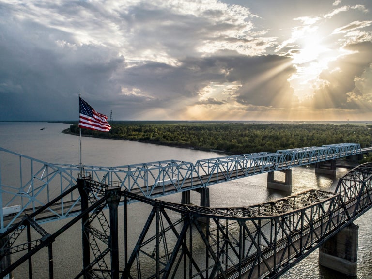 Vicksburg Bridge over the Mississippi River with greenery shown and sun shining in the background as the American flag flies. 