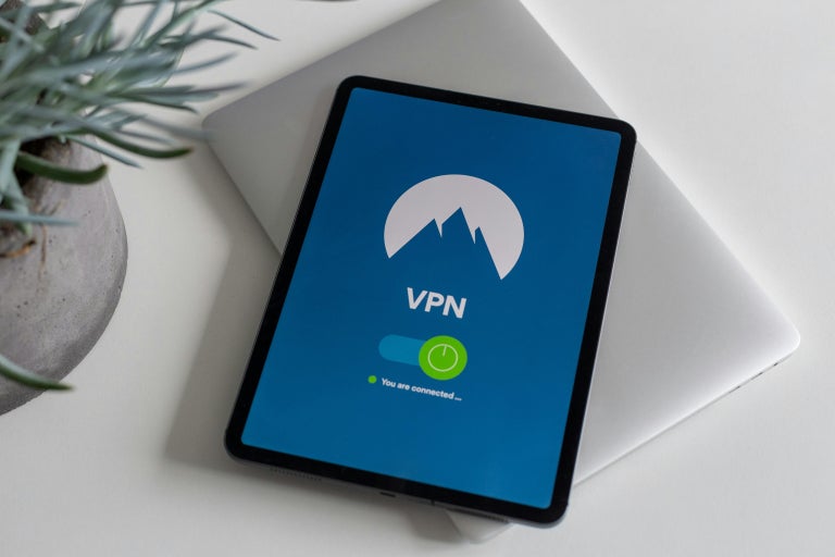 Tablet sitting on top of a laptop computer next to a houseplant displaying “VPN” with a logo of a mountain in the background.