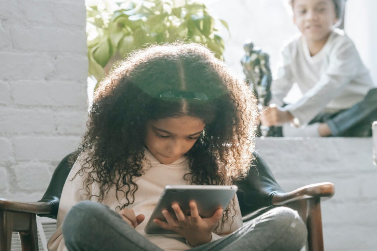 Young girl looking down at a tablet while young boy in the background plays with a toy