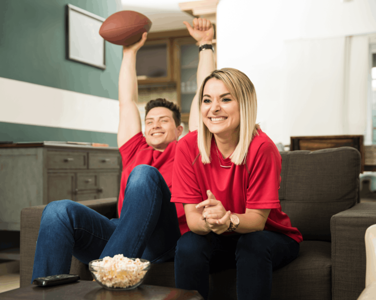 Couple watching football on a couch; man celebrating with hands raised with football in hand